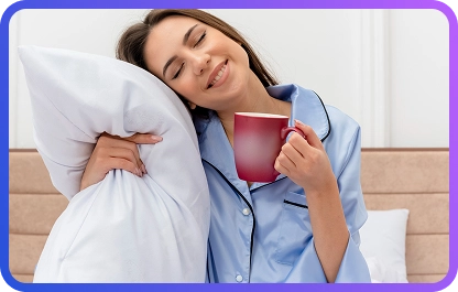 Zrelaksowana kobieta w niebieskiej piżamie, trzymająca poduszkę i czerwony kubek, ciesząca się spokojnym porankiem w miękkiej, przytulnej sypialni.